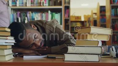 非裔美国人英俊的年轻<strong>男学生</strong>正坐在桌子旁，图书馆里有一大堆书。 他的头在躺着